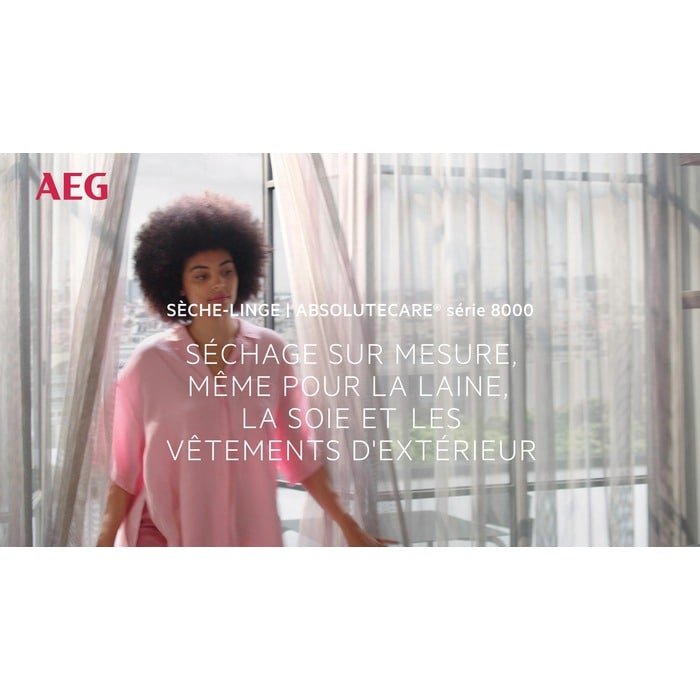 Une femme vêtue d'un haut rose se tient près de grandes fenêtres aux rideaux diaphanes ; un texte en français souligne la technologie avancée du sèche-linge AEG TR81A3G6BOE.