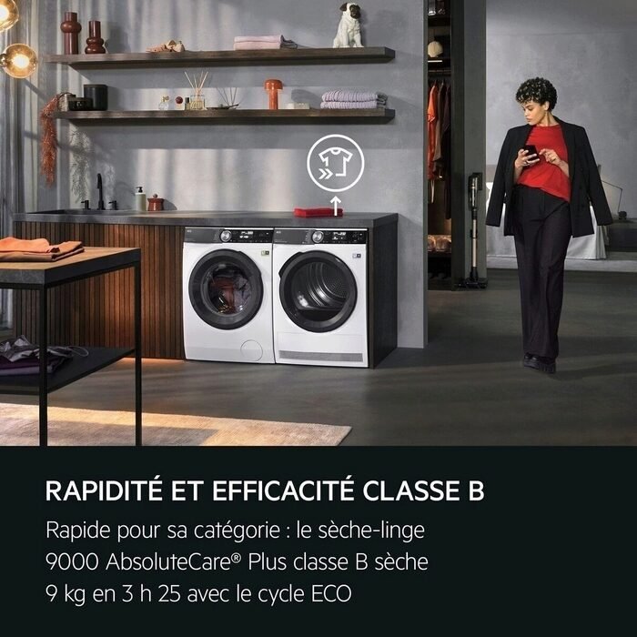 Une femme se tient dans une buanderie moderne à côté d'un lave-linge et d'un sèche-linge AEG TR95A9B6BW, tout en consultant son téléphone.