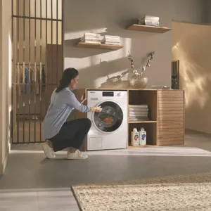 Une femme est agenouillée dans une buanderie, chargeant des vêtements dans un lave-linge MIELE WWB360WCS à chargement frontal.