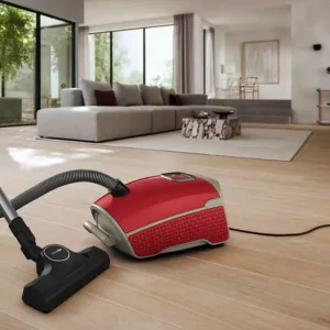 Un aspirateur-traîneau Miele GUARD L1 RED PULSE est placé sur un sol en bois clair dans un salon moderne avec de grandes fenêtres.