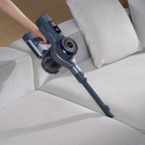 Une main utilise un aspirateur sans fil NARWAL pour enlever les miettes dans les interstices d'un canapé gris clair.
