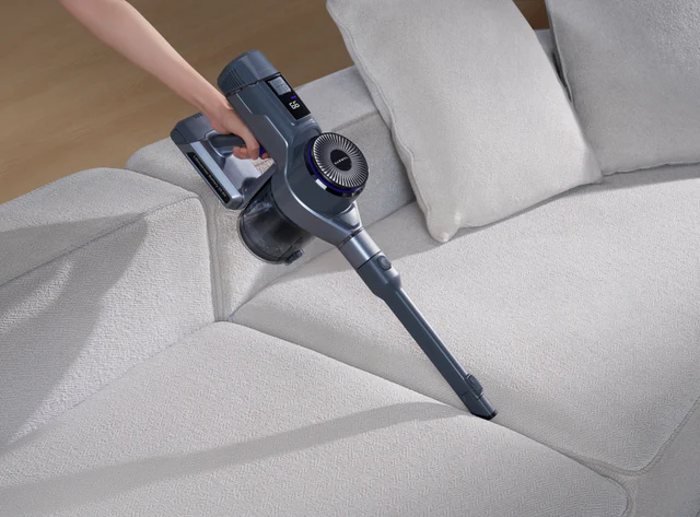 Une main utilise un aspirateur sans fil NARWAL pour enlever les miettes dans les interstices d'un canapé gris clair.