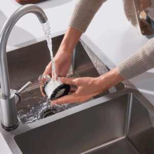 Une personne lave une brosse à récurer NARWAL à l'eau courante dans un évier de cuisine moderne en acier inoxydable.