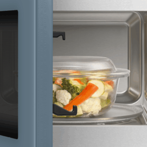 Un récipient en verre contenant des légumes mélangés est placé à l'intérieur d'un four à micro-ondes ouvert en acier inoxydable, à côté d'une élégante machine à café SMEG MOC01BLMEU.