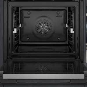 Le four encastrable SIEMENS HB974GLB1 avec une porte en verre noir est ouvert et vide à côté d'une étagère remplie de bocaux, de bouteilles et d'un livre de cuisine.