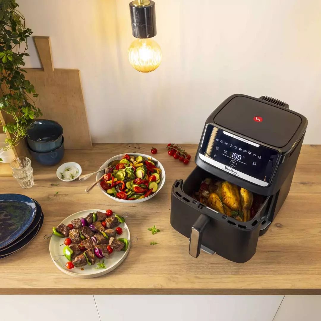 La friteuse MOULINEX EZ8428F0 est posée sur le comptoir avec du poulet rôti à l'intérieur, entourée d'assiettes de brochettes grillées et de salades fraîches, démontrant ainsi sa polyvalence dans la préparation de repas délicieux.