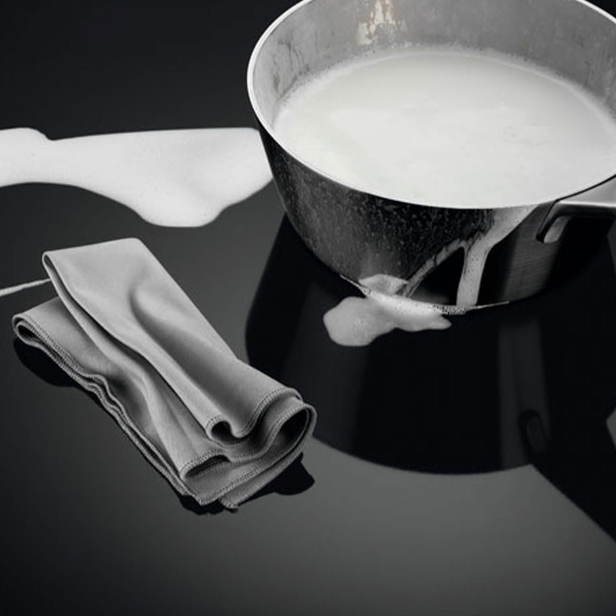 Le lait bout sur la plaque de cuisson à induction AEG IKE64471FB noire et élégante, avec un chiffon plié à proximité.