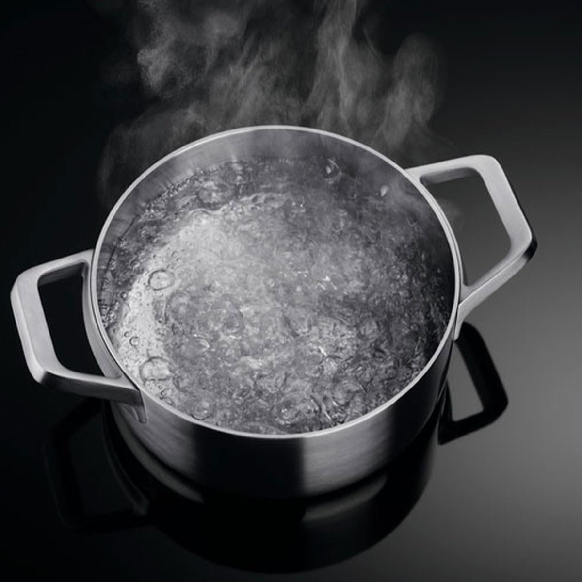 Une casserole d'eau en métal bout et la vapeur monte, posée sur une surface sombre au-dessus d'une plaque à induction AEG IKE64471FB.