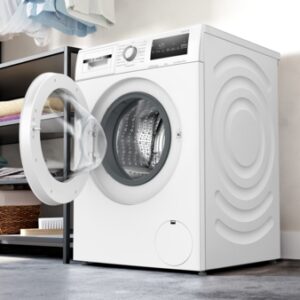 Le lave-linge à chargement frontal BOSCH WAN2827AFR est présenté avec sa porte ouverte dans une buanderie moderne dotée d'étagères et de paniers.