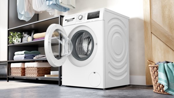 Le lave-linge à chargement frontal BOSCH WAN2827AFR est présenté avec sa porte ouverte dans une buanderie moderne dotée d'étagères et de paniers.