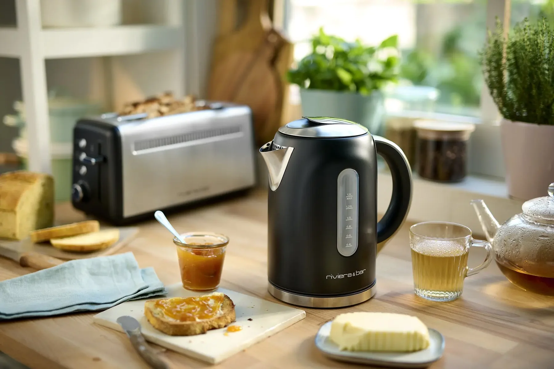 La bouilloire électrique noire BBT577 (Copie) de RIVIERA & BAR est posée sur le comptoir de la cuisine avec des toasts, du beurre, de la confiture, du thé et un grille-pain - parfait pour une routine matinale élégante.
