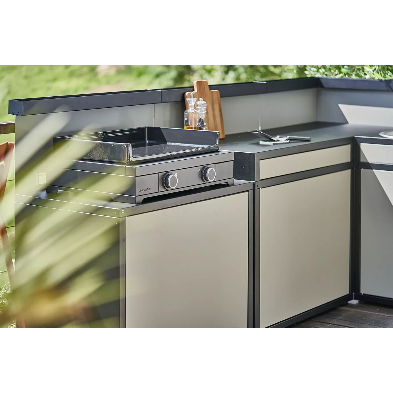 Cuisine extérieure moderne comprenant un gril en acier inoxydable, une plancha FORGE ADOUR MODERN E60 AN, des armoires élégantes et un comptoir avec des planches à découper et des condiments.