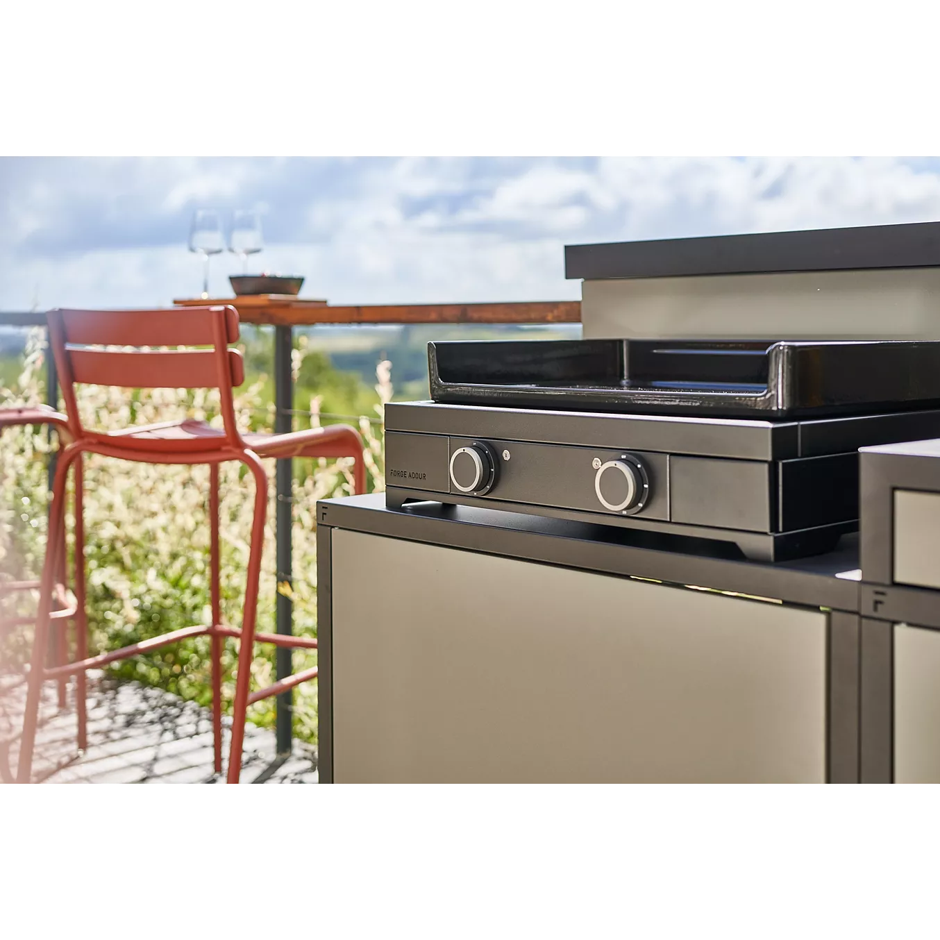 Gril en plein air sur un patio avec une plancha FORGE ADOUR MODERN E60 AN, avec un tabouret de bar rouge et des verres à vin sur une table en arrière-plan.