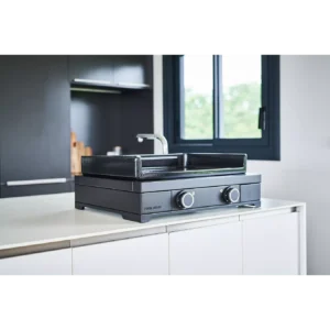 A FORGE ADOUR MODERN E60 UNE plancha avec deux boutons est posée sur un comptoir de cuisine blanc près d'une fenêtre, apportant style et fonctionnalité à l'espace moderne.