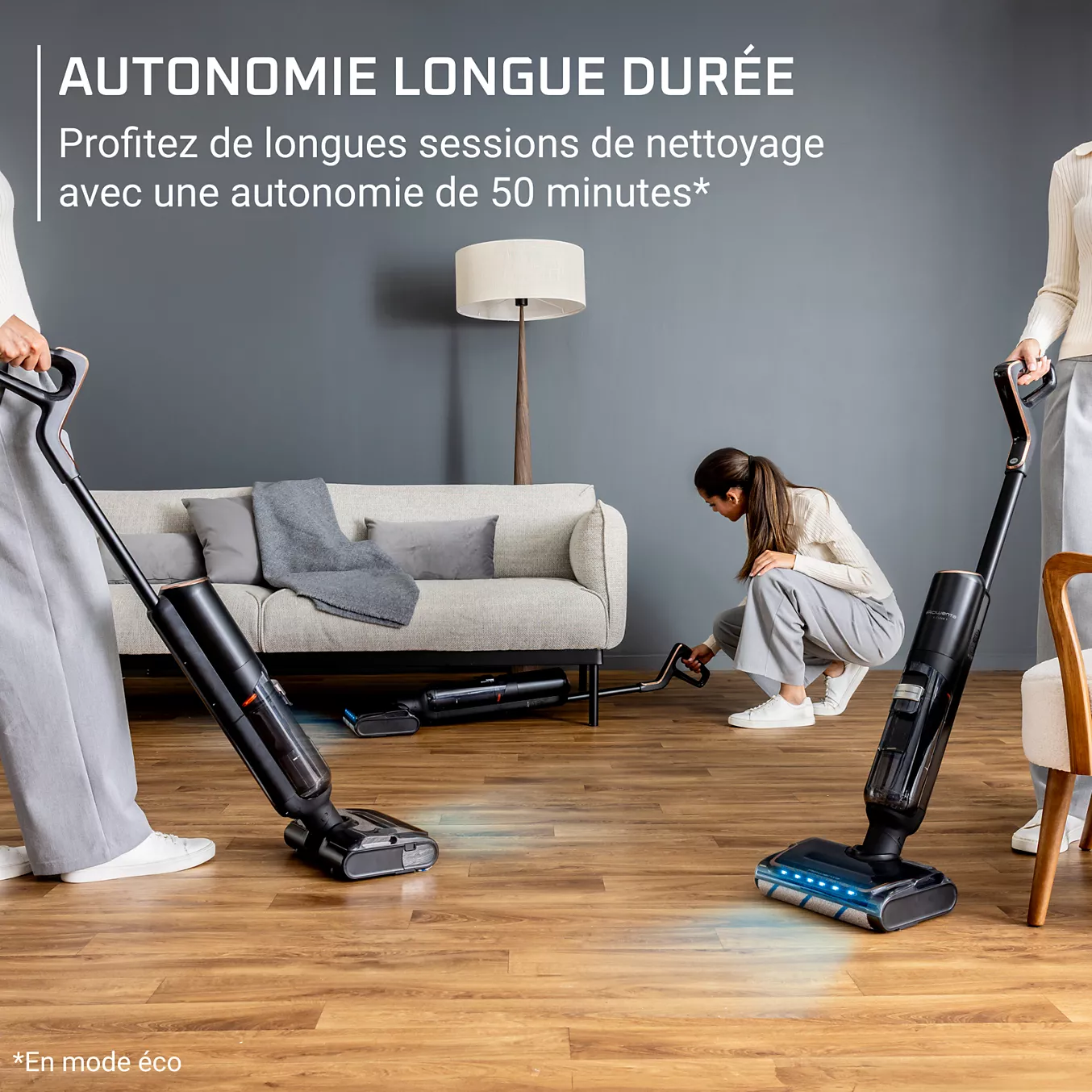 Deux personnes utilisent l'aspirateur sans fil ROWENTA GZ5736E0 pour nettoyer un salon composé d'un canapé, d'une lampe et d'un sol en bois.