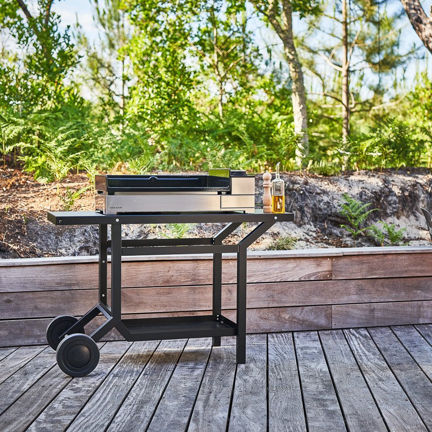 Un chariot moderne de grillades en plein air FORGE ADOUR TRA_N avec des condiments est posé sur une terrasse en bois, entourée d'arbres et de verdure luxuriante.