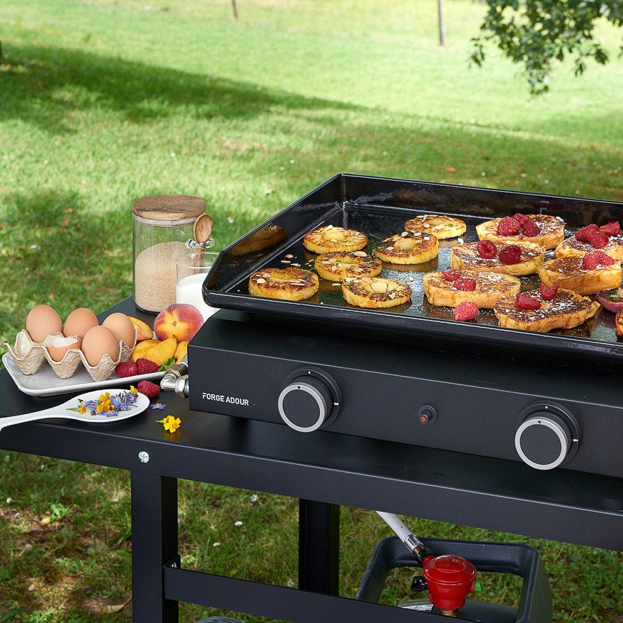 Cuisson de pain perdu aux fruits sur une plaque FORGE ADOUR TRA_N en plein air ; les œufs et les ingrédients sont disposés sur la table dans une cour herbeuse.