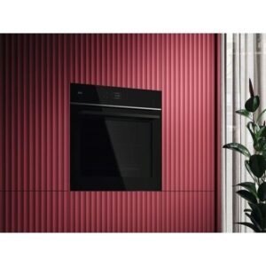 Le four AEG noir encastré est placé dans un mur rouge nervuré à côté d'une fenêtre avec un rideau blanc et une plante verte, créant un look élégant qui s'associe parfaitement avec des appareils modernes comme le sèche-linge AEG TX7PB631SB.