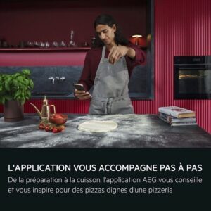 Portant un tablier, une personne consulte son téléphone tout en préparant de la pâte à pizza sur le comptoir de la cuisine, tandis que l'AEG TX7PB631SB fonctionne silencieusement en arrière-plan.