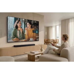 Détendez-vous sur votre canapé avec la télécommande SAMSUNG TMR55R85H, en appréciant une scène où deux femmes discutent et dînent sur un grand téléviseur mural.