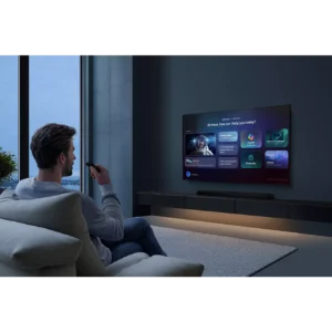 Un homme utilise une télécommande pour interagir avec son téléviseur intelligent SAMSUNG TMR65R85H, qui affiche diverses icônes d'application alors qu'il est assis sur son canapé.