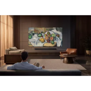 Un homme se détend dans un salon moderne, en regardant une peinture de fruits affichée sur un grand écran SAMSUNG TMR65R95H fixé au mur.
