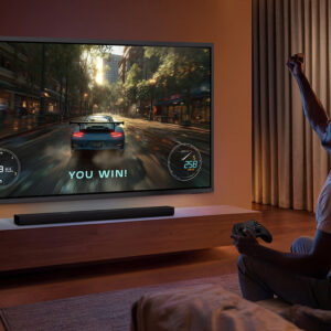 Une personne célèbre sa victoire dans un jeu vidéo de course automobile diffusé sur le grand téléviseur SAMSUNG TQ83S95H, dans un salon confortable.