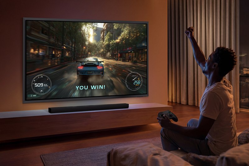 Une personne célèbre sa victoire dans un jeu vidéo de course automobile diffusé sur le grand téléviseur SAMSUNG TQ83S95H, dans un salon confortable.
