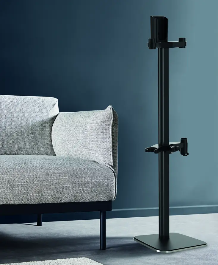 Dans une pièce moderne aux murs et à la moquette bleus, un canapé gris minimaliste côtoie une ROWENTA DOCKING STATION 7 aux lignes épurées et un grand support de sol noir.