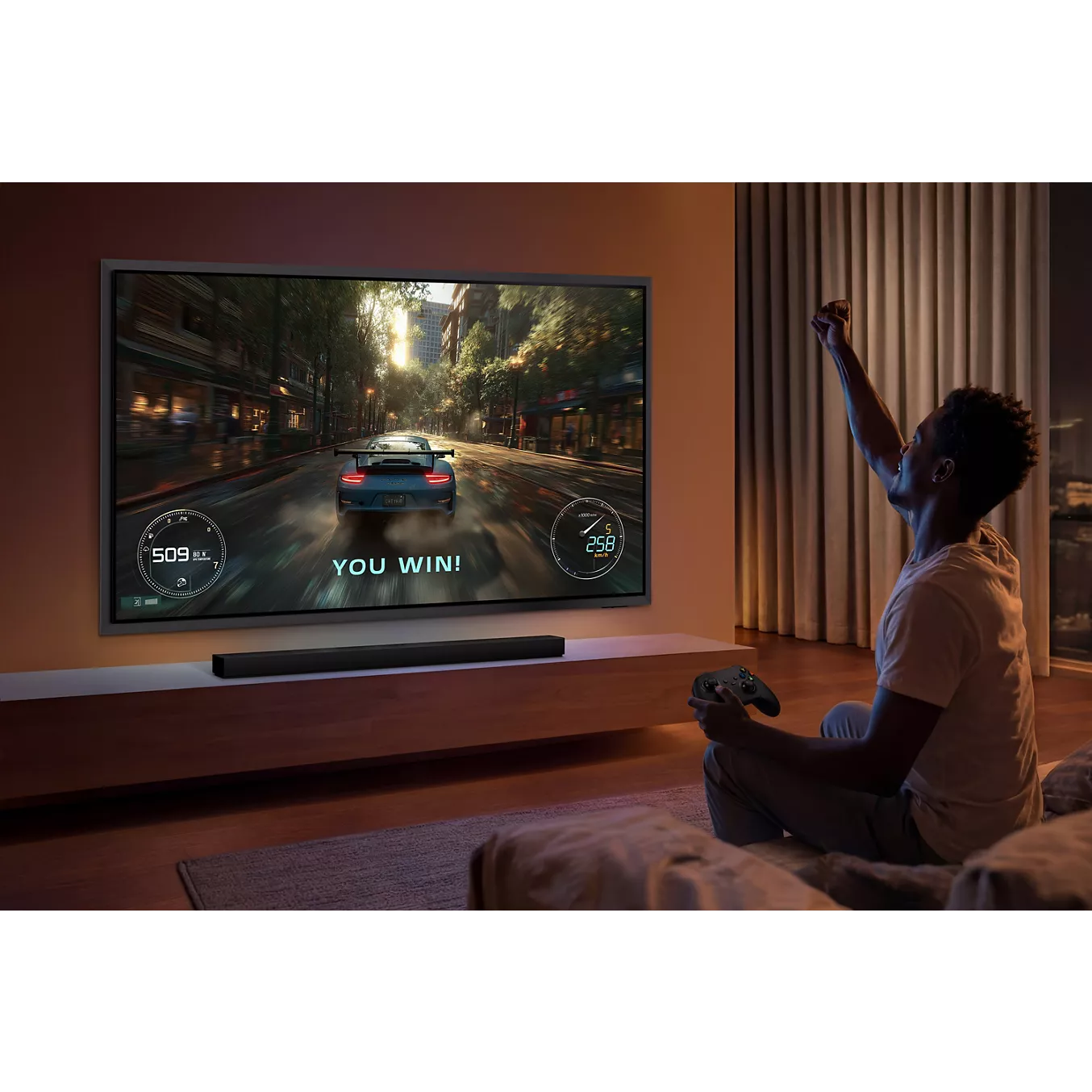 Une personne fait la fête sur son canapé après avoir gagné un jeu vidéo de course automobile sur un grand écran de télévision SAMSUNG TQ65S95H (Copie).