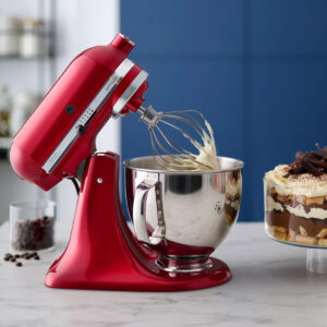 Un robot pâtissier KITCHENAID 5KSM175PSEFL rouge, dont le fouet est recouvert de pâte, trône sur un plan de travail à côté d'un trifle.
