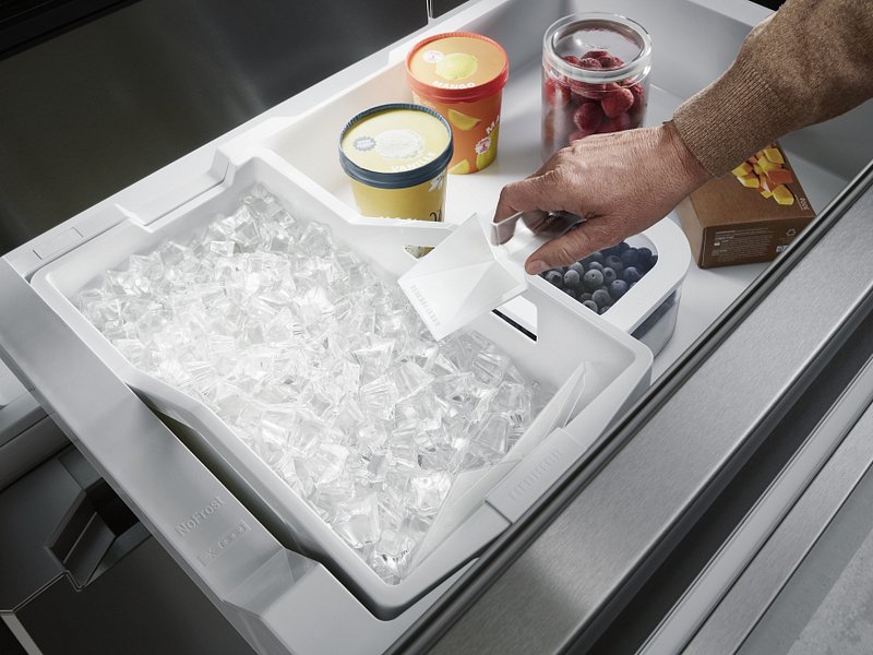 On voit ici quelqu'un prélever à la main des glaçons dans le tiroir congélateur du modèle LIEBHERR MBSDDI9524-20, à côté duquel se trouvent de la glace, des fruits rouges et une boîte de surgelés.