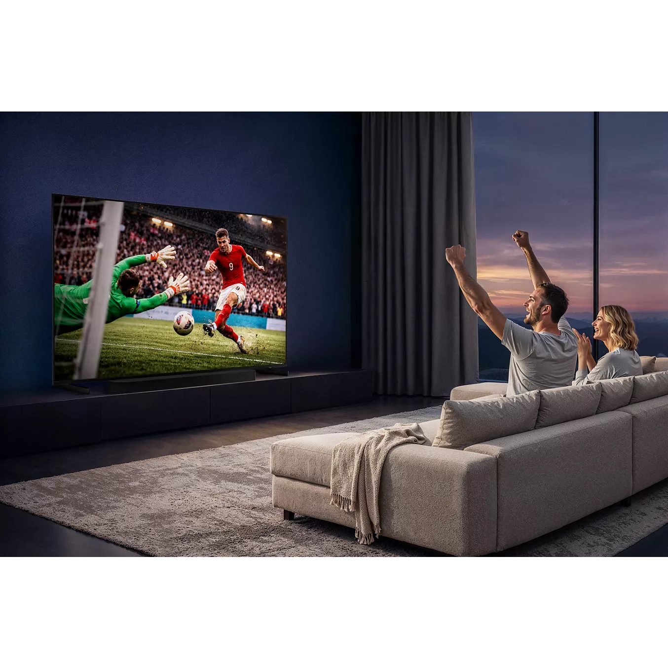 Un couple applaudit, confortablement installé sur un canapé, tout en regardant un match de football sur le téléviseur SAMSUNG TMR75R85H dans un salon moderne.