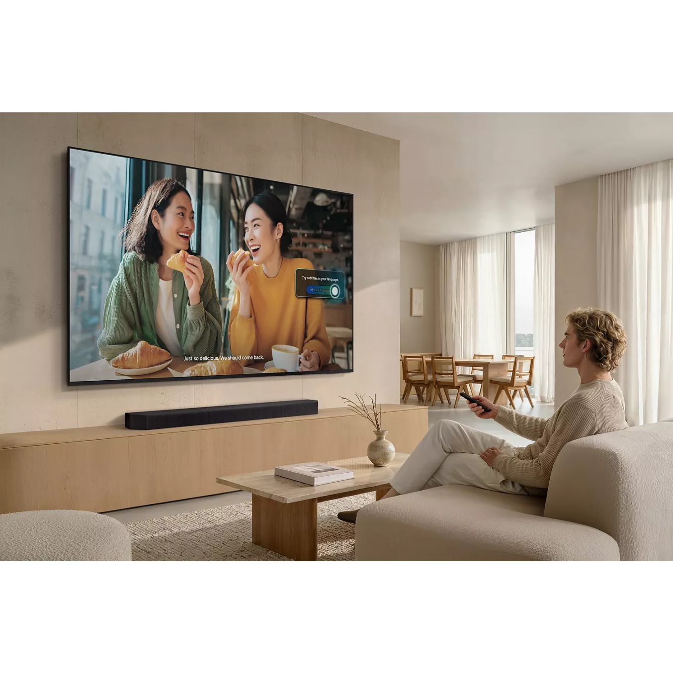 Une personne assise sur un canapé utilise une télécommande pour regarder la télévision, où l'on voit deux femmes souriantes en train de manger ensemble sur l'élégant téléviseur SAMSUNG TMR75R85H.