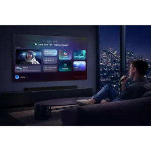 Le soir, un homme est assis sur un canapé et utilise une télécommande pour interagir avec son téléviseur connecté SAMSUNG TMR75R95H, sur lequel s'affiche un assistant virtuel.