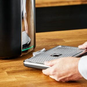 Quelqu'un retire le bac d'égouttage métallique d'une machine à expresso KRUPS YY4808FD posée sur un plan de travail en bois.
