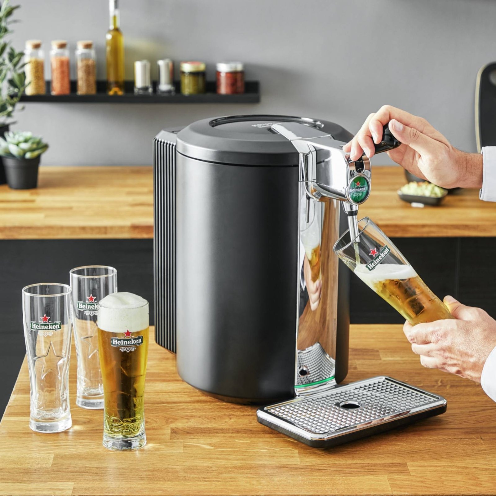 Quelqu'un verse de la bière à partir d'une tireuse KRUPS YY4808FD dans un verre posé sur un plan de travail en bois, à côté de deux verres déjà remplis.