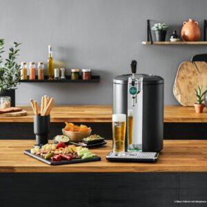 Un robinet à bière domestique, accompagné d'un verre de bière, de snacks et de légumes, trône sur un plan de travail en bois, à côté de l'élégante machine à café KRUPS YY4808FD, au milieu de plantes et d'étagères.
