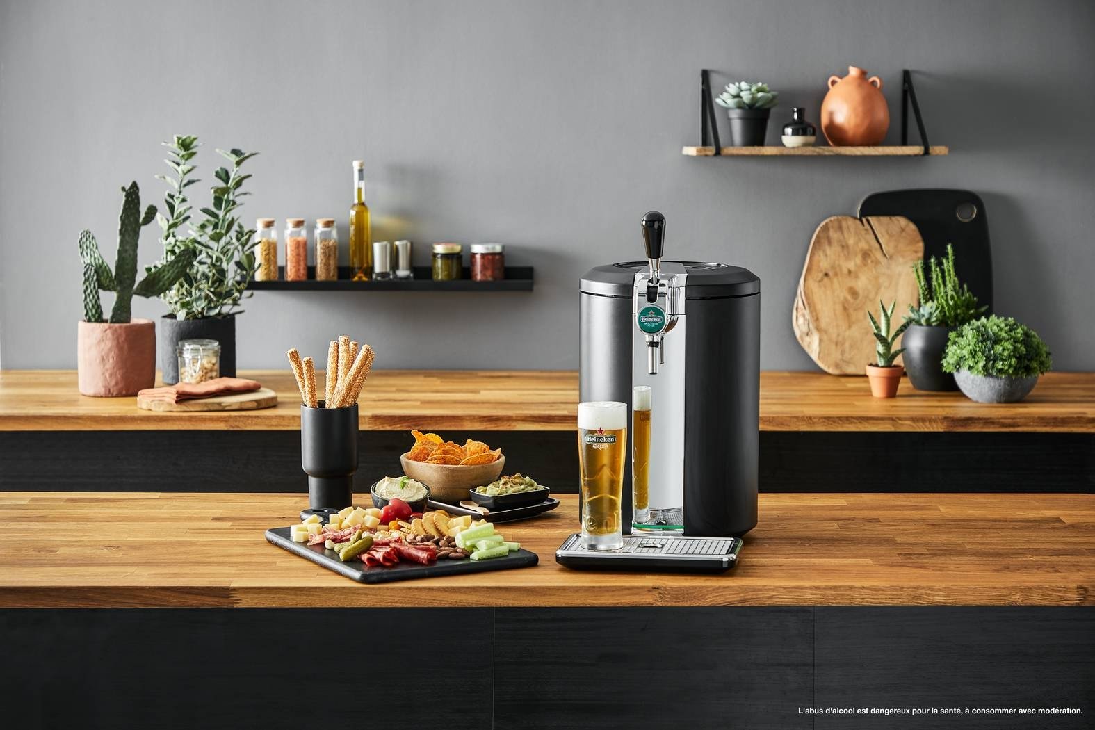 Un robinet à bière domestique, accompagné d'un verre de bière, de snacks et de légumes, trône sur un plan de travail en bois, à côté de l'élégante machine à café KRUPS YY4808FD, au milieu de plantes et d'étagères.