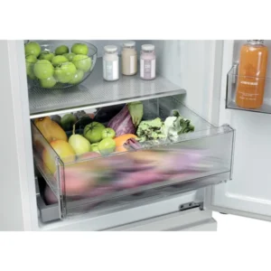 Ouvrez le réfrigérateur HAIER HTR3518ENMW : vous y trouverez un bac à légumes rempli de légumes frais, des pommes sur une étagère et des bouteilles dans le compartiment de la porte.