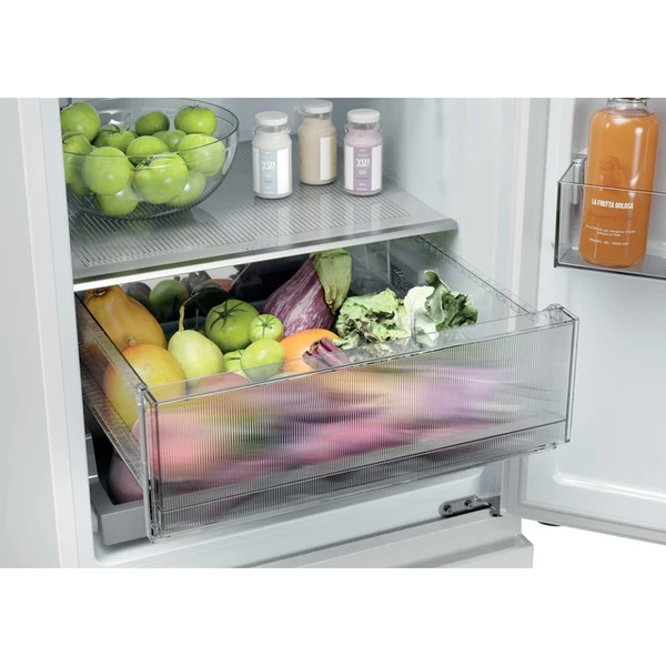 Ouvrez le réfrigérateur HAIER HTR3518ENMW : vous y trouverez un bac à légumes rempli de légumes frais, des pommes sur une étagère et des bouteilles dans le compartiment de la porte.