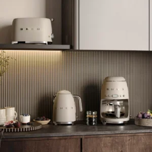 Le modèle SMEG TSF01CHMEU (Copier), composé d'un grille-pain, d'une bouilloire et d'une cafetière de couleur beige, est présenté sur un plan de travail de cuisine, accompagné de tasses, de fruits et d'objets décoratifs.