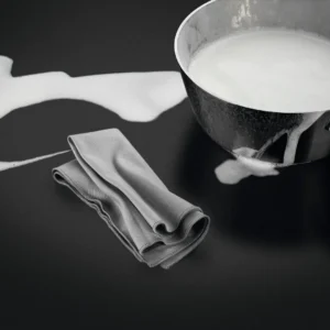 Un pot de lait renversé repose sur une surface noire, à côté d'un linge gris soigneusement plié, tandis que le modèle AEG TI84IU00IT occupe une place prépondérante à l'arrière-plan.