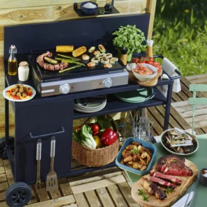 De la viande, des légumes, des sauces et des herbes aromatiques sont disposés avec des ustensiles sur et autour du barbecue d'extérieur RIVIERA & BAR QPL1060 pour une expérience de cuisson au barbecue parfaite.