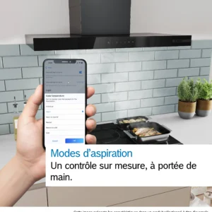 Une main tient un téléphone qui commande la hotte de cuisine DWB97DN, située au-dessus d'une cuisinière sur laquelle se trouvent une casserole et une poêle. Un texte en français s'affiche à l'écran.