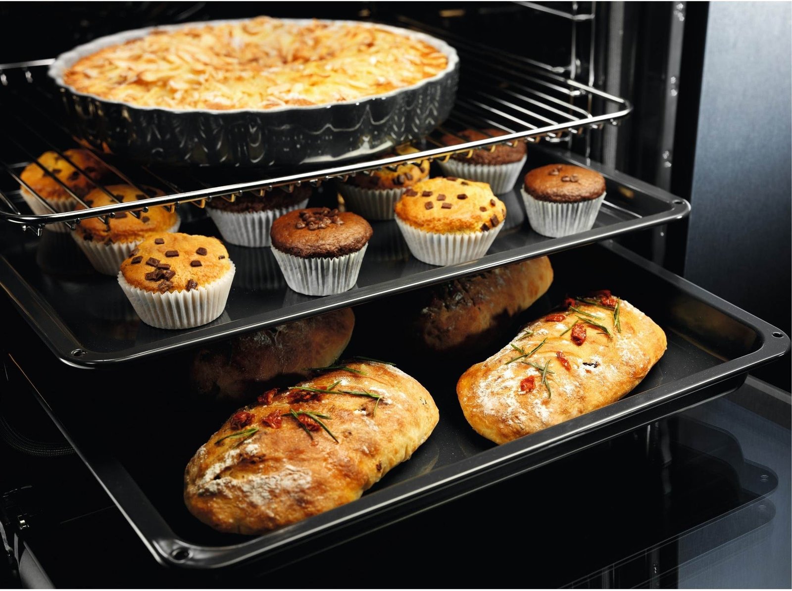 Trois grilles de four, sur lesquelles sont disposés une tarte, des muffins et des miches de pain, sont en train de cuire dans le four moderne ELECTROLUX EOF3C40TH.