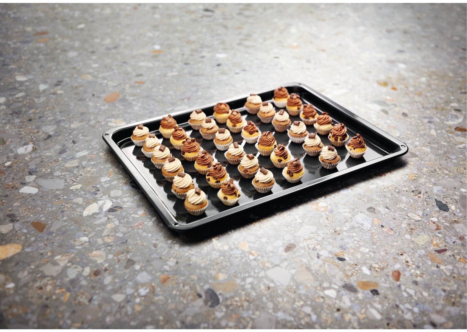 Une plaque de cuisson sur laquelle sont disposées des rangées de mini-cupcakes assortis, tout juste sortis du four ELECTROLUX EOF3C40TH, repose sur un plan de travail moucheté.