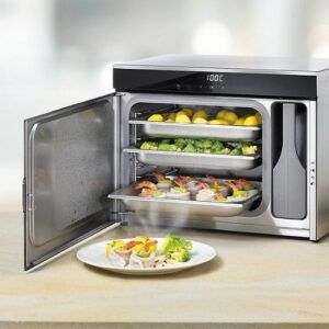 Un four à vapeur MIELE DG6019NR contenant des plateaux de plats, avec une assiette de plats cuits posée sur le plan de travail devant.