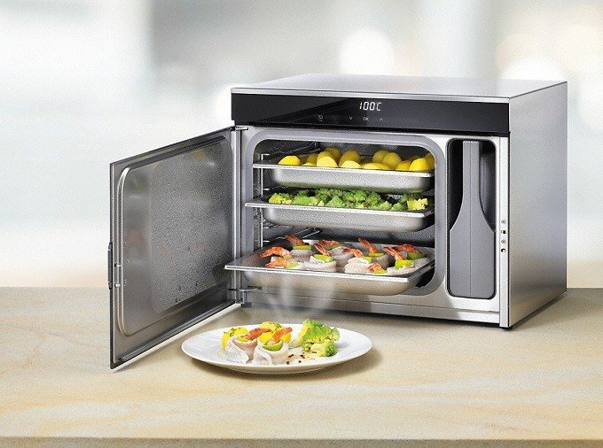 Un four à vapeur MIELE DG6019NR contenant des plateaux de plats, avec une assiette de plats cuits posée sur le plan de travail devant.