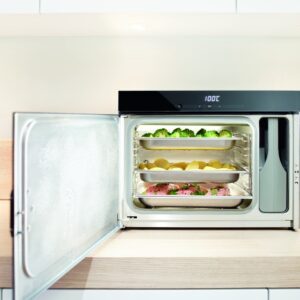 Le four à vapeur MIELE DG6019NR est posé, porte ouverte, sur le plan de travail d'une cuisine moderne ; à l'intérieur se trouvent des plateaux de brocolis, de pommes de terre et de poulet assaisonné.